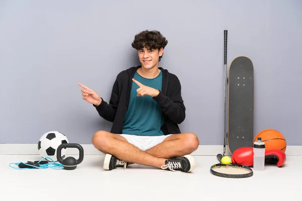 Young sport man sitting on the floor around many sport elements pointing finger to the side