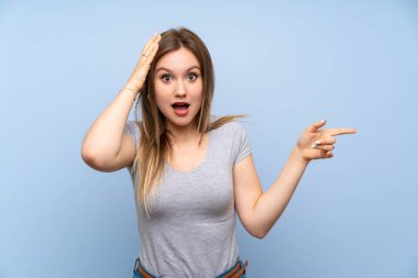 İzole mavi duvar üzerinde genç kız sürpriz ve tarafına parmak işaret