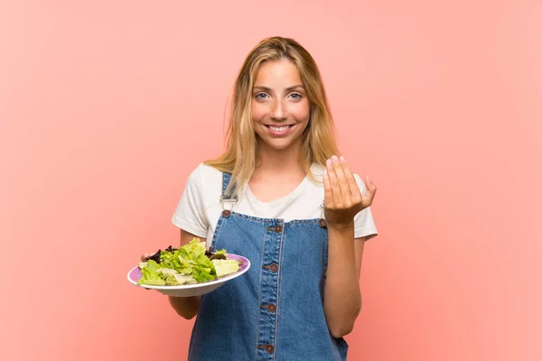 Blonde young woman with salad over isolated pink wall inviting to come with hand. Happy that you came