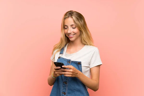Mujer joven rubia feliz con un teléfono móvil sobre una pared rosa aislada —  Fotos de Stock