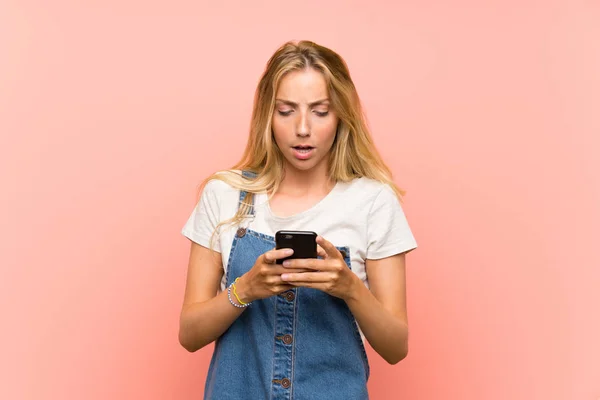 Mujer joven rubia con un teléfono móvil sobre una pared rosa aislada —  Fotos de Stock