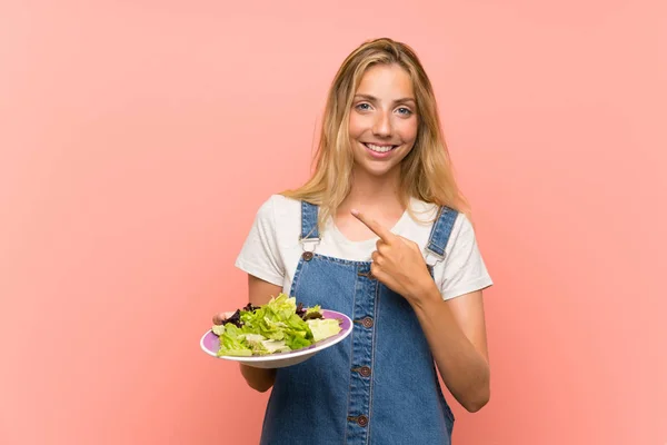 Blonde young woman with salad over isolated pink wall pointing to the side to present a product