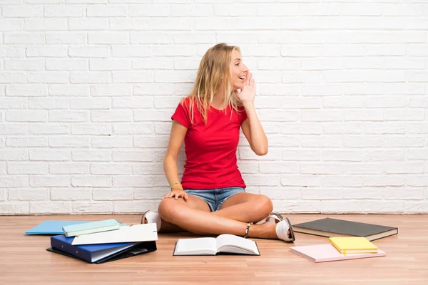 Young blonde student girl with many books on the floor shouting with mouth wide open