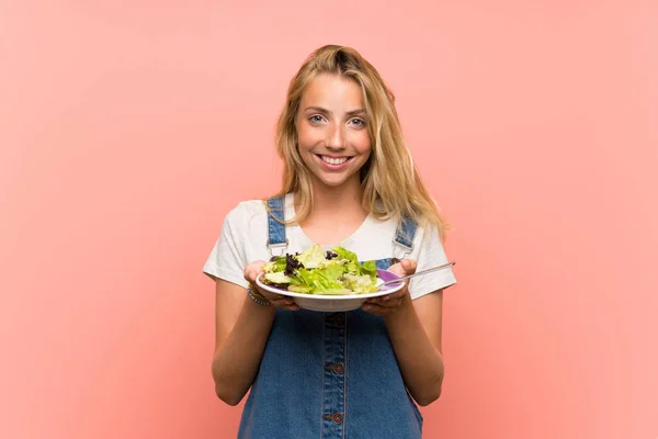 Happy Blonde young woman with salad over isolated pink wall