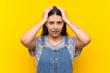 Sürpriz yüz ifadesi ile izole sarı arka plan üzerinde dungarees genç kadın
