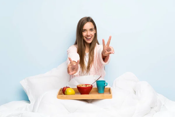 Young woman in dressing gown with breakfast smiling and showing victory sign