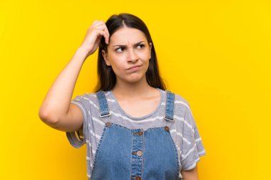 Izole sarı arka plan üzerinde dungarees genç kadın şüpheleri olan ve yüz ifadesi karıştırmayın