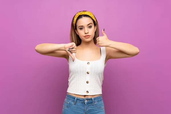 Pretty young woman over isolated purple wall making good-bad sign. Undecided between yes or not