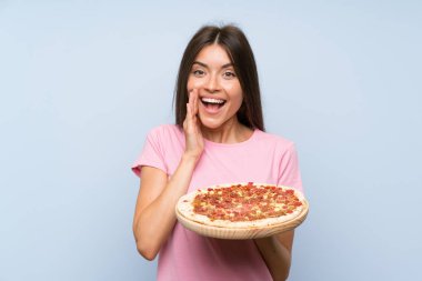 Sürpriz ve şok yüz ifadesi ile izole mavi duvar üzerinde bir pizza tutan Pretty genç kız