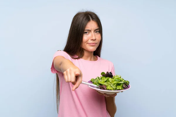 Young woman with salad over isolated blue background points finger at you with a confident expression