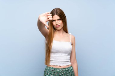 İzole edilmiş mavi arka planda genç bir kadın baş parmağını negatif ifadeyle aşağı gösteriyor.