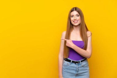 İzole edilmiş sarı arka planda genç bir kadın bir ürün sunmak için kenarı işaret ediyor.