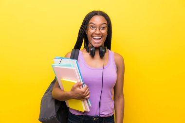 Sürpriz yüz ifadesi ile izole sarı duvar üzerinde uzun örgülü saçlı Afro-Amerikan genç öğrenci kız