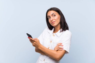 İzole mavi duvar üzerinde bir cep telefonu ile genç kadın omuzkaldırma sırasında şüphe jest yapma