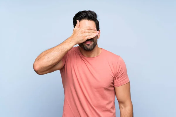 Handsome young man in pink shirt over isolated blue background covering eyes by hands. Do not want to see something