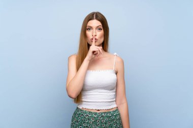 İzole edilmiş mavi arka planda genç bir kadın sessizlik işareti gösteriyor. Parmağını ağzına sok.