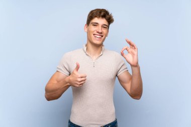 Izole mavi duvar üzerinde yakışıklı genç adam ok işareti ve başparmak yukarı jest gösteren