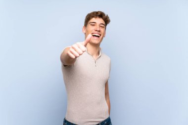 Yakışıklı genç adam izole mavi duvar üzerinde başparmak yukarı çünkü iyi bir şey oldu