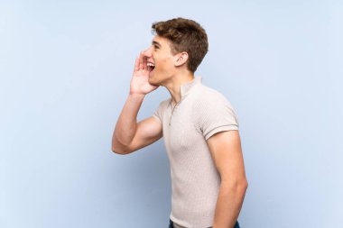 Izole mavi duvar üzerinde yakışıklı genç adam yanal açık ağız ile bağırarak