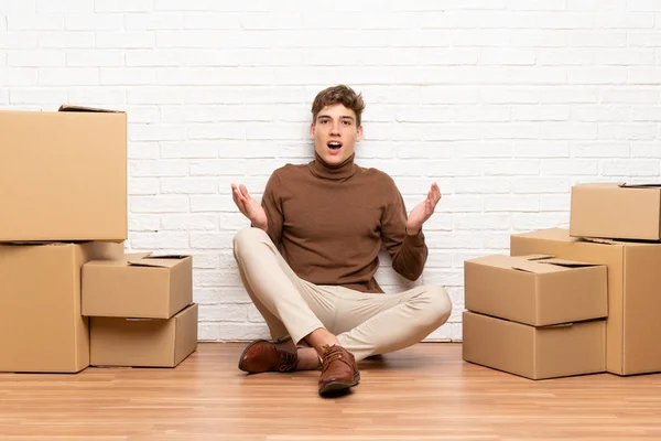 Handsome young man moving in new home among boxes with surprise facial expression