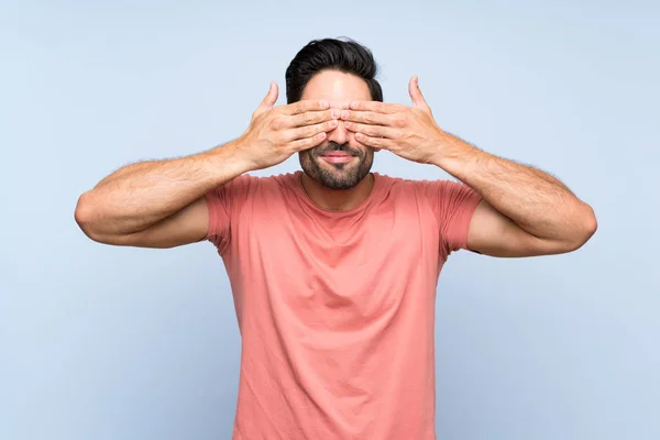 Handsome young man in pink shirt over isolated blue background covering eyes by hands