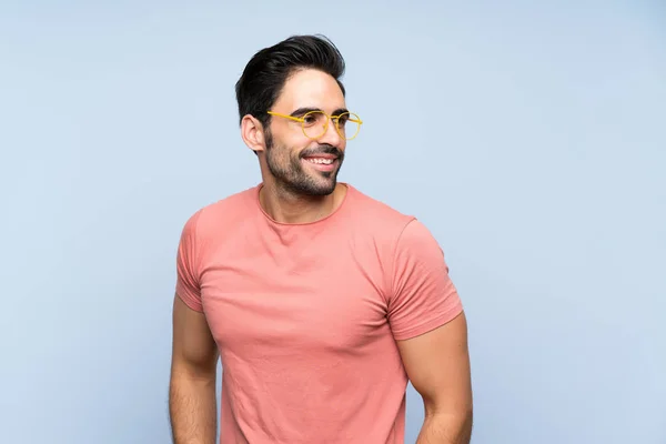 メガネと笑顔で孤立した青い背景の上にピンクのシャツのハンサムな若者 — ストック写真