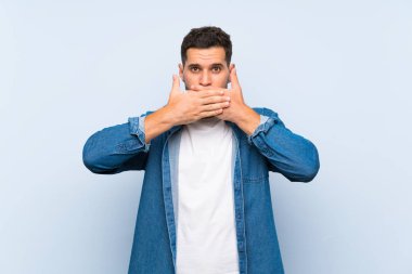 İzole edilmiş mavi arka planda yakışıklı bir adam elleriyle ağzını kapatıyor.
