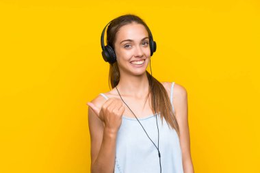 Bir ürün sunmak için yan işaret izole sarı duvar üzerinde müzik dinleyen genç kadın