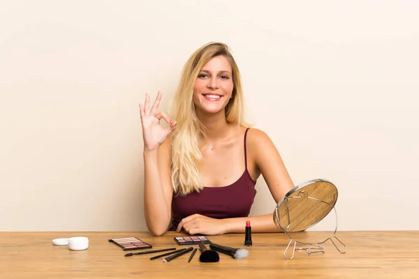 年轻的金发女人与化妆品在桌子上显示一个确定的迹象与手指 — 图库照片