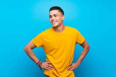 Izole mavi arka plan üzerinde sarı gömlekli genç adam kalça kolları ile poz ve gülümseyerek