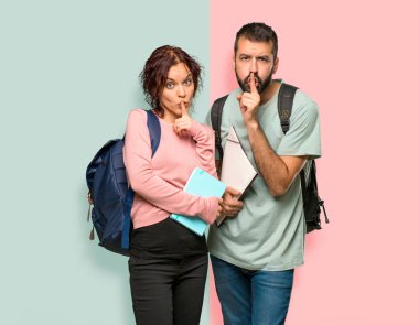 Sırt çantaları ve renkli duvara parmak koyarak sessizlik jest iişareti gösteren kitap ile iki öğrenci