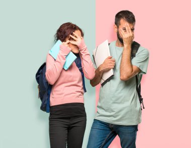 Sırt çantaları ve kitapları ile iki öğrenci elleriyle gözleri kaplayan ve renkli duvarda parmaklarından bakarak