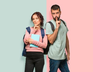 Sırt çantaları ve renkli duvarda ağız ve sessizlik jest kapanış bir işaret gösteren kitap ile iki öğrenci