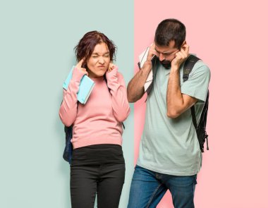 Sırt çantaları ve kulakları elleriyle kaplayan kitapları olan iki öğrenci. Renkli duvarda sinirli ifade