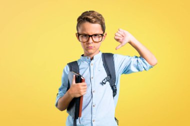 Sırt çantası ve gözlük sarı arka plan üzerinde olumsuz ifade ile başparmak aşağı işareti gösteren öğrenci çocuk