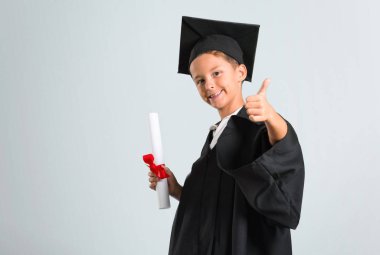 Küçük çocuk jest kadar bir başparmak veren ve gri arka plan üzerinde gülümseyen mezun