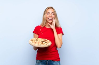 Genç sarışın kadın sürpriz ve şok yüz ifadesi ile izole mavi duvar üzerinde bir pizza tutan