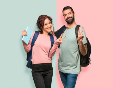 Sırt çantaları ve kitapları ile iki öğrenci gülümseyerek ve renkli duvarda iki eliyle zafer işareti gösteren