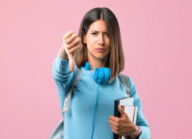 Mavi kazak ve kulaklık pembe arka plan üzerinde olumsuz ifade ile başparmak aşağı işareti gösteren genç öğrenci kız