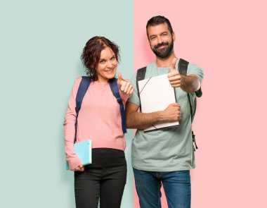 Sırt çantaları ve kitapları olan iki öğrenci başparmak kadar jest veren ve renkli duvarda gülümseyerek