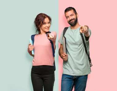 Sırt çantaları ve kitapları olan iki öğrenci, renkli duvarda kendine güvenen bir ifadeyle sizi işaret ediyor