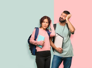 Sırt çantaları ve kitapları olan iki öğrenci, şüpheleri olan ve renkli duvara kafa kaşırken yüzünü karıştıran