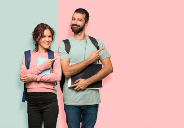 Renkli duvarda bir ürün sunmak için bir parmak ile yan işaret sırt çantaları ve kitapları ile iki öğrenci