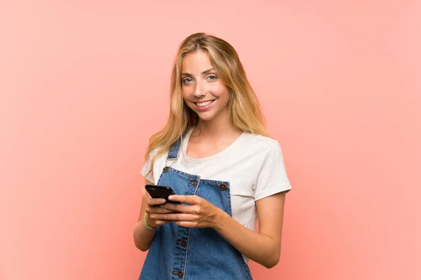 Mujer joven rubia feliz con un teléfono móvil sobre una pared rosa aislada —  Fotos de Stock