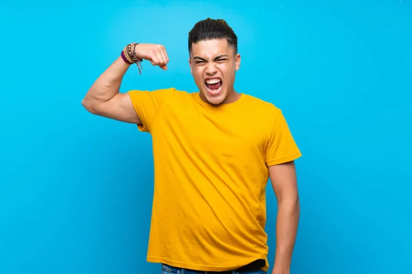Young man with yellow shirt over isolated blue background doing strong gesture