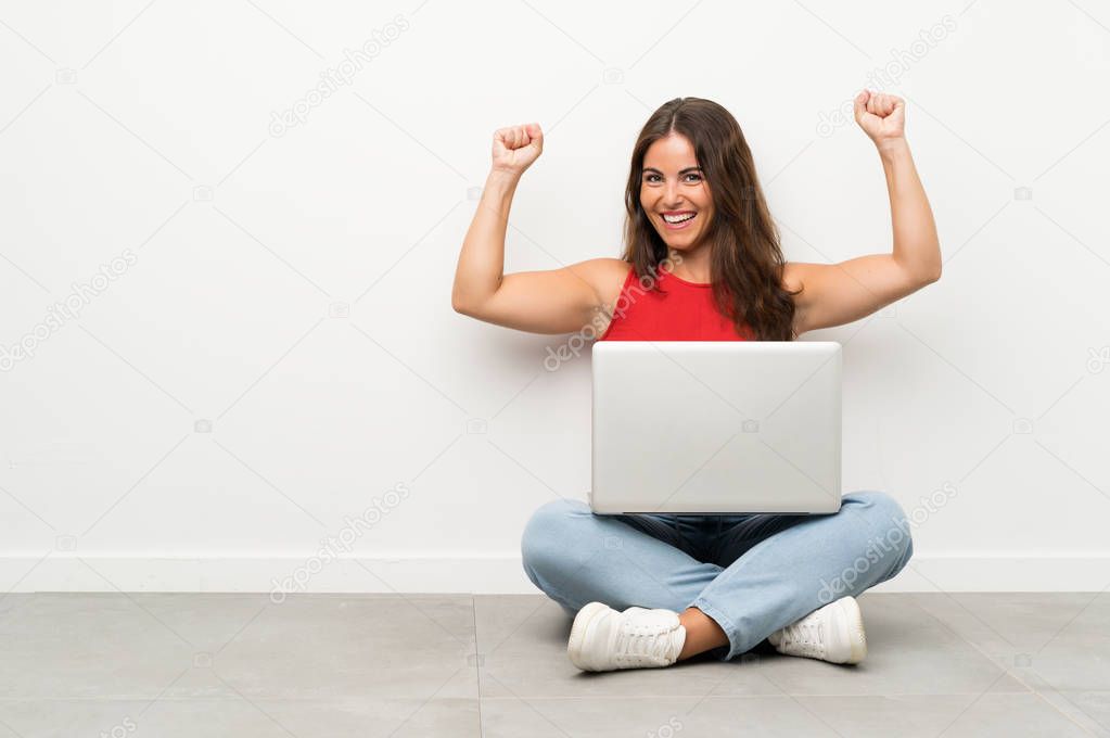 Young woman with a laptop sitting on the floor celebrating a victory