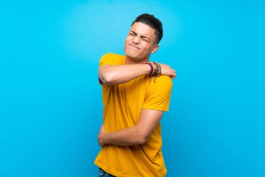 Izole mavi arka plan üzerinde sarı gömlekli genç adam bir çaba için omuz ağrısı muzdarip