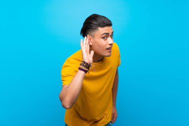 Izole mavi arka plan üzerinde sarı gömlekli genç adam kulağına el koyarak bir şey dinlerken