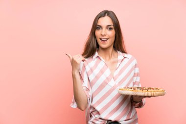 Genç kadın izole pembe duvar üzerinde bir ürün sunmak için yan işaret bir pizza tutan