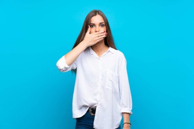 İzole edilmiş mavi arka planı olan genç bir kadın elleriyle ağzını kapatıyor.